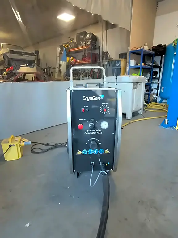 A dry ice blaster on wheels standing on a workshop floor, its hose running behind it.